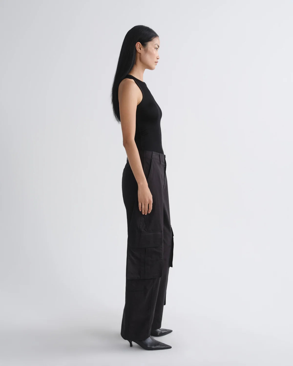A woman in a black sleeveless top and black wide-leg cargo pants stands in profile against a light gray studio background.