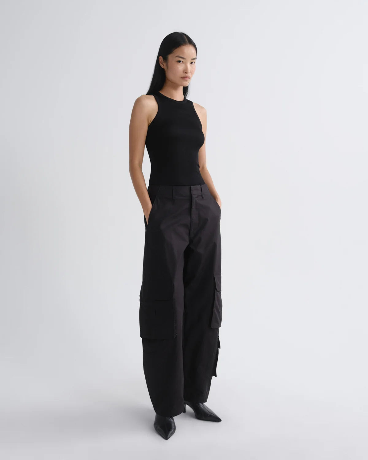 A model stands in a studio wearing a black sleeveless top and black wide-leg cargo pants with black pointed shoes against a light gray background.