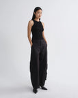 A model stands in a studio wearing a black sleeveless top and black wide-leg cargo pants with black pointed shoes against a light gray background.