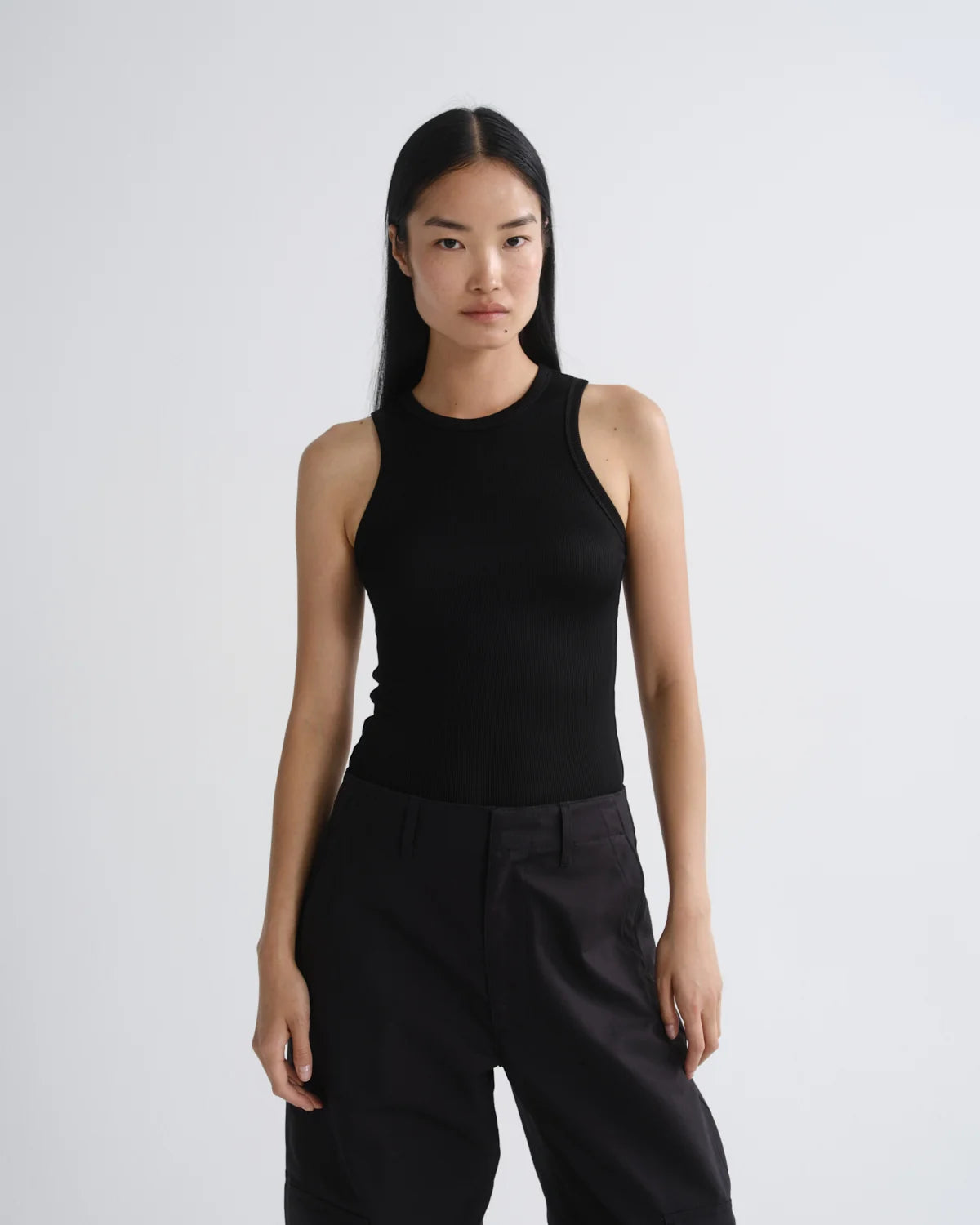 A model wearing a black sleeveless ribbed tank top and black wide-leg trousers, standing against a light gray studio background.