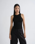 A model wearing a black sleeveless ribbed tank top and black wide-leg trousers, standing against a light gray studio background.