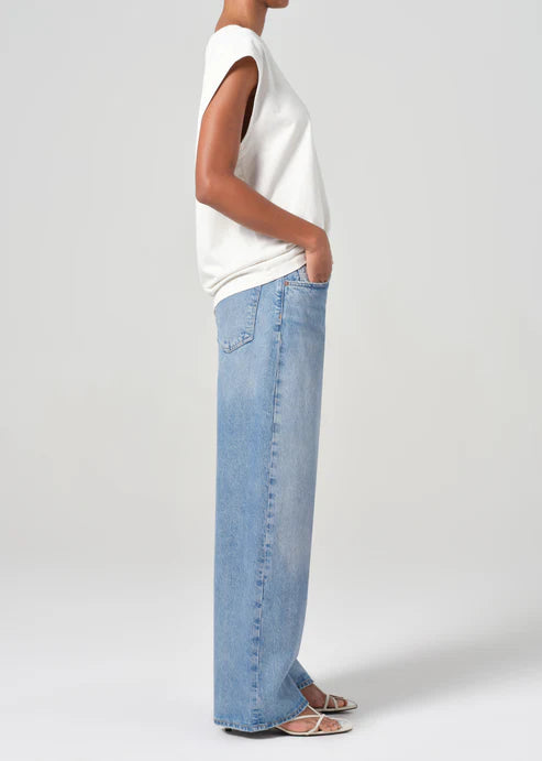 Person wearing a white sleeveless top and light blue jeans on a plain background side view