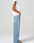 Person wearing a white sleeveless top and light blue jeans on a plain background side view