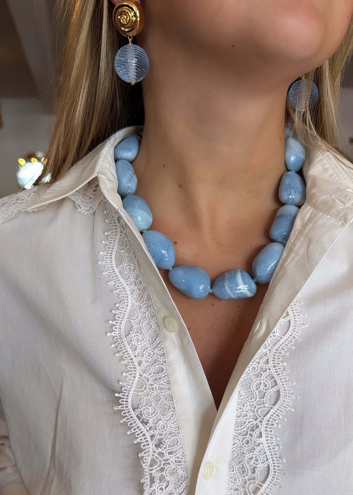 A close-up of a model wearing a chunky light blue stone necklace and matching drop earrings, paired with a white lace-trimmed button-up shirt.