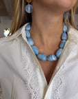 A close-up of a model wearing a chunky light blue stone necklace and matching drop earrings, paired with a white lace-trimmed button-up shirt.