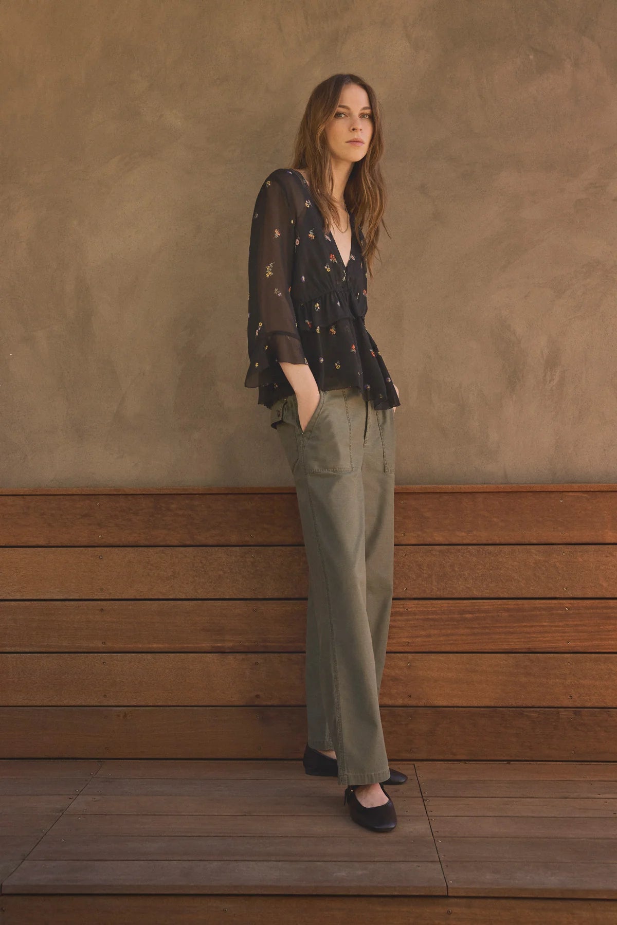 A woman stands against a textured brown wall and wooden bench, wearing a sheer black long-sleeve blouse with small gold floral motifs, olive green wide-leg trousers, and black flats.