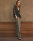 A woman stands against a textured brown wall and wooden bench, wearing a sheer black long-sleeve blouse with small gold floral motifs, olive green wide-leg trousers, and black flats.