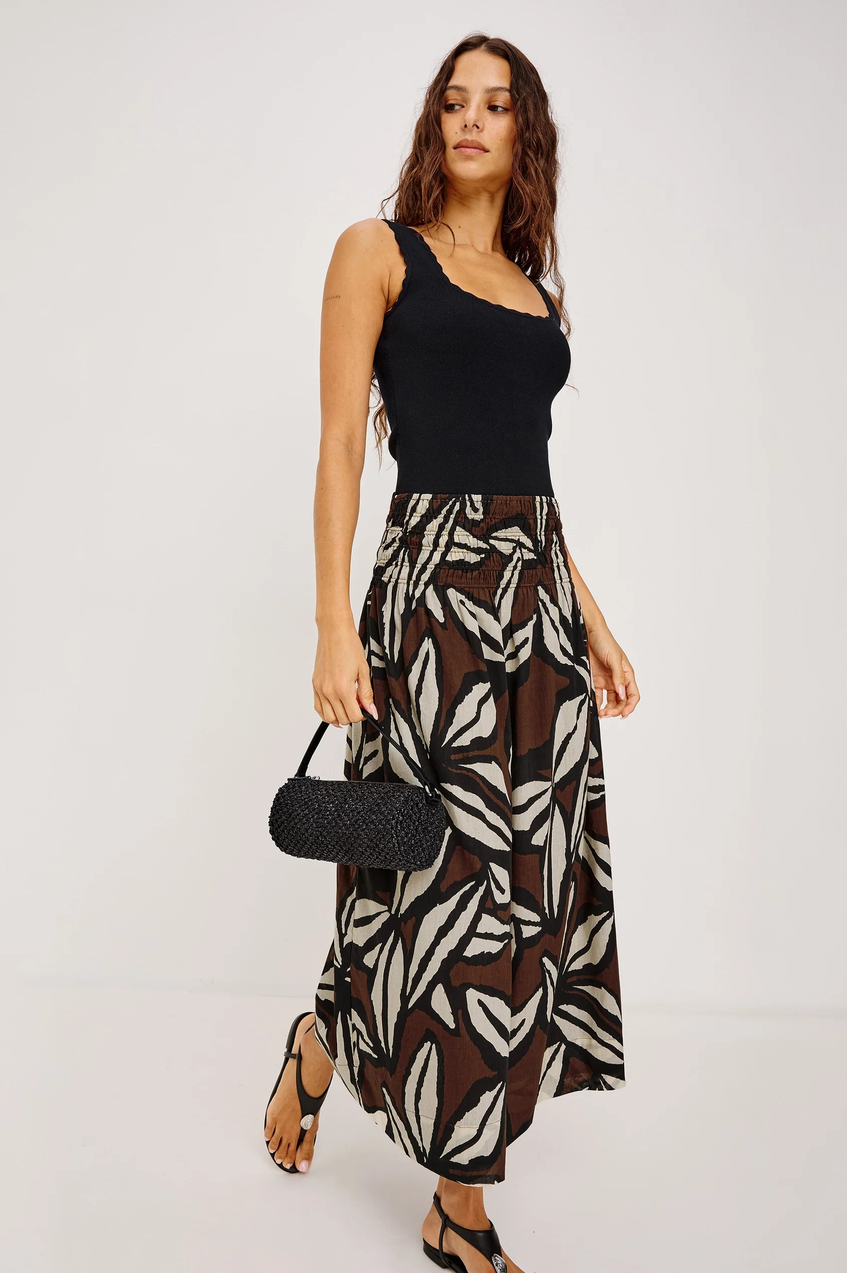 Woman wearing a black top and patterned skirt holding a black handbag on a white background