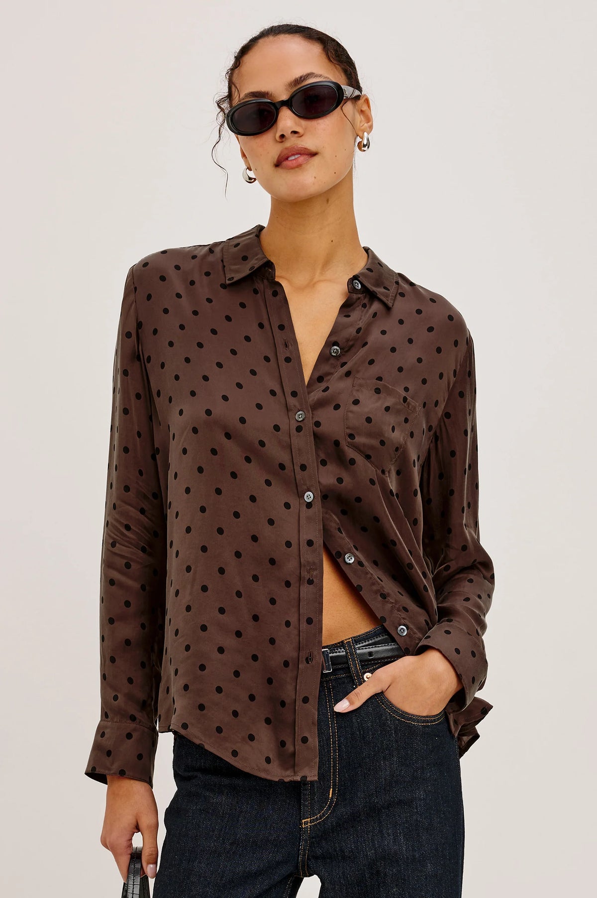 A woman modeling a brown long-sleeve button-up shirt with black polka dots, paired with dark blue jeans and black sunglasses, against a plain light gray background.