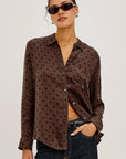 A woman modeling a brown long-sleeve button-up shirt with black polka dots, paired with dark blue jeans and black sunglasses, against a plain light gray background.