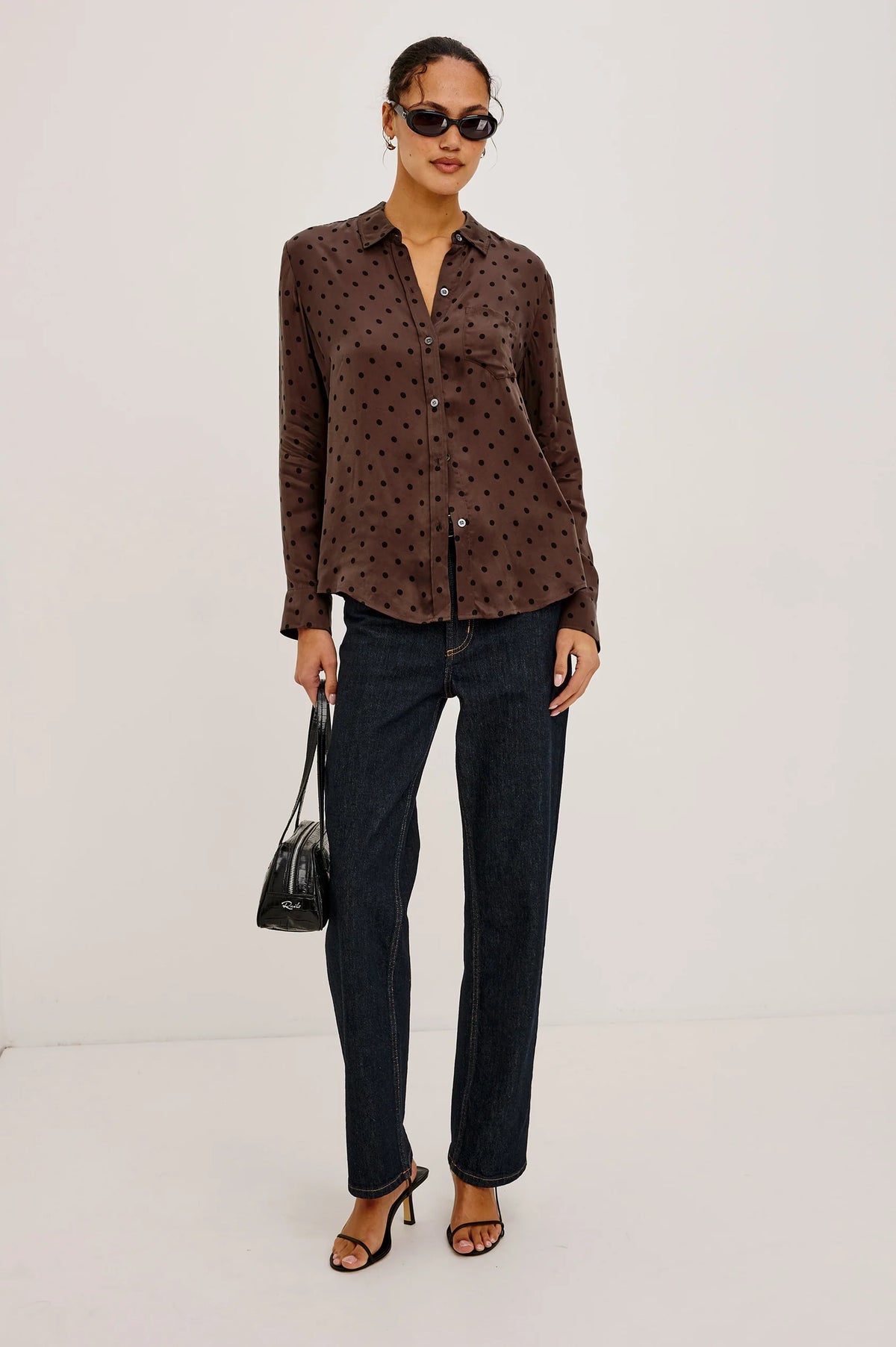 A woman modeling a brown long-sleeve button-up shirt with a small black polka dot pattern, paired with dark blue jeans, black sunglasses, and black heeled sandals, holding a black handbag against a plain white studio background.