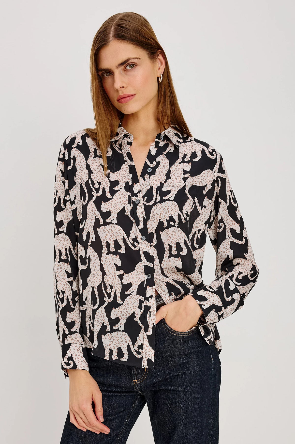 A woman modeling a black long-sleeve button-up blouse with an allover beige animal print, paired with dark blue jeans, against a plain light gray studio background.