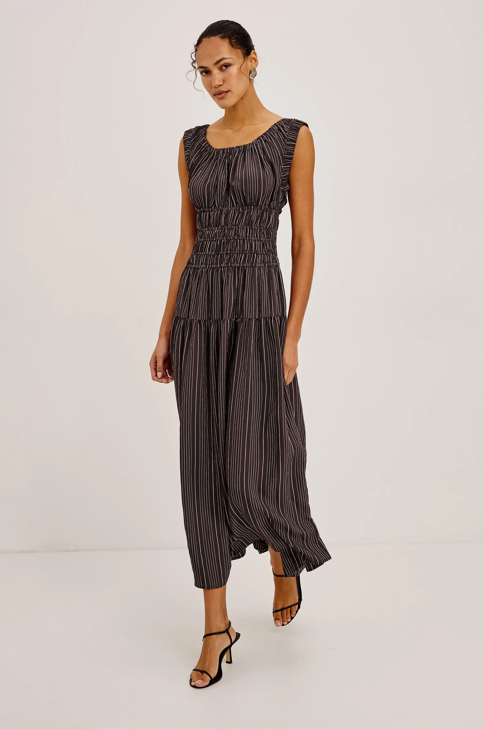 A woman modeling a sleeveless dark gray and black vertically striped jumpsuit with a smocked waist and pleated skirt, paired with black strappy heels, against a plain light gray studio background.