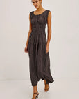 A woman modeling a sleeveless dark gray and black vertically striped jumpsuit with a smocked waist and pleated skirt, paired with black strappy heels, against a plain light gray studio background.