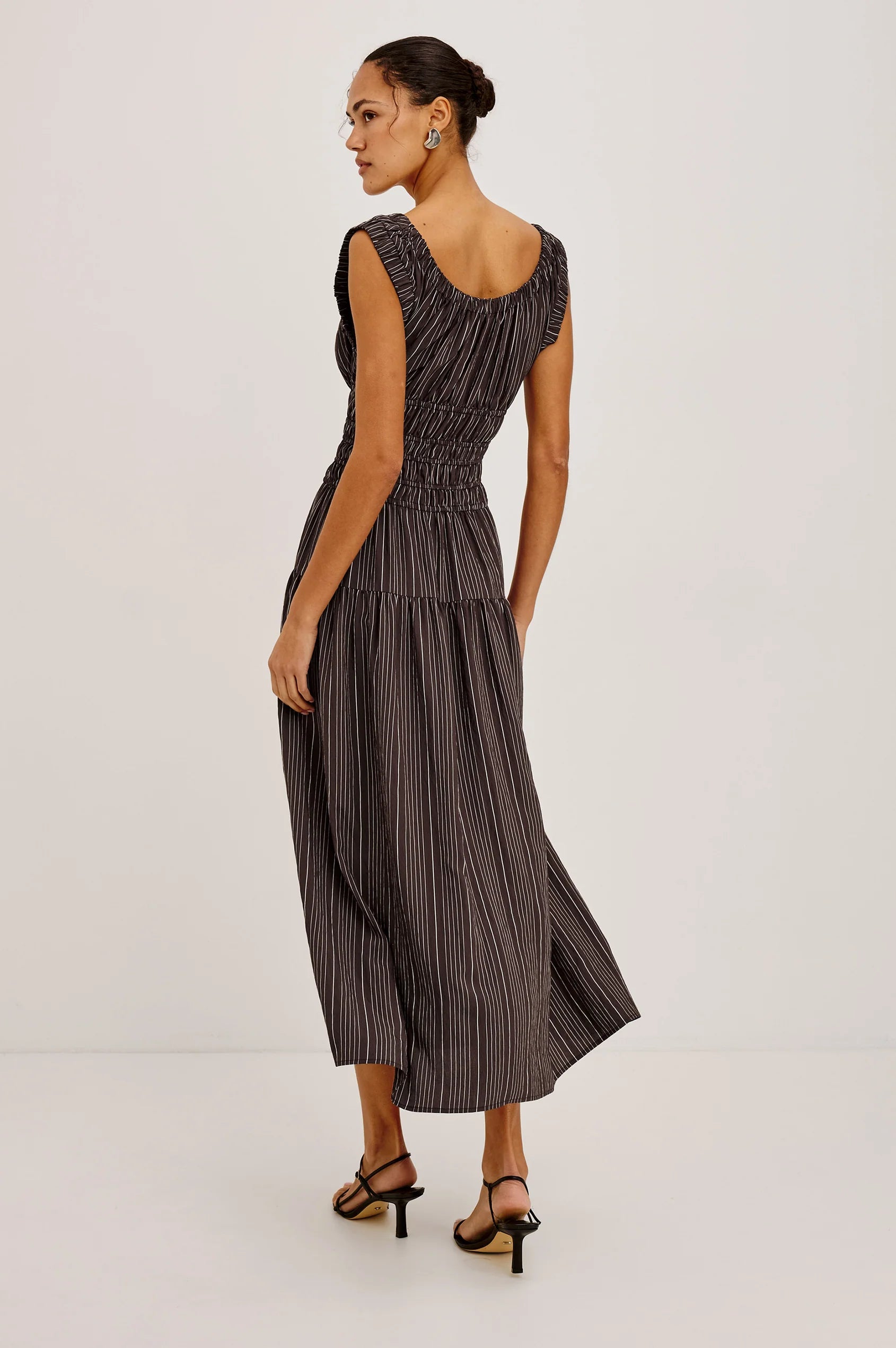 A woman modeling a sleeveless midi dress with vertical pinstripes in a dark gray and black colorway, shown from a three-quarter back angle against a plain white studio background.