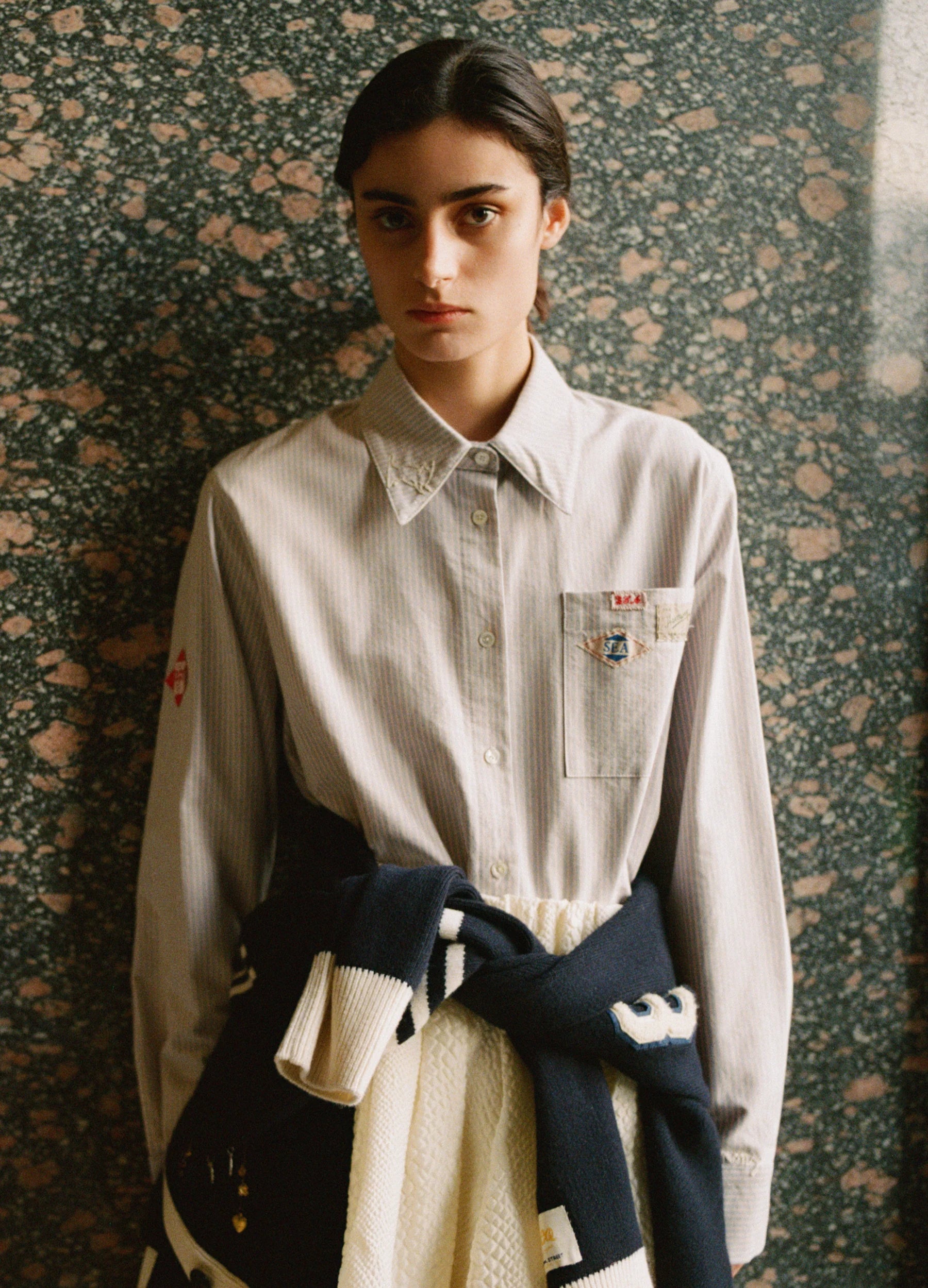 Person wearing a light-colored shirt with embroidered details against a textured wall.