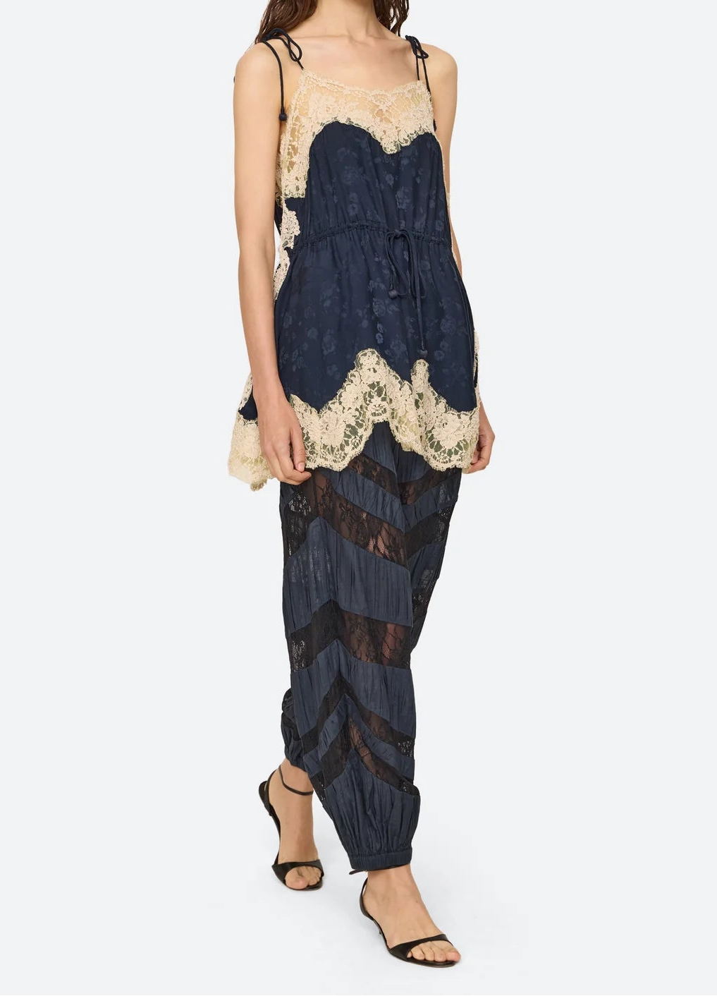 A model wearing a dark navy blue lace-trimmed camisole top and matching pleated harem pants with sheer chevron detailing, paired with black strappy heels, against a light gray studio background.