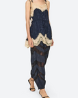 A model wearing a dark navy blue lace-trimmed camisole top and matching pleated harem pants with sheer chevron detailing, paired with black strappy heels, against a light gray studio background.