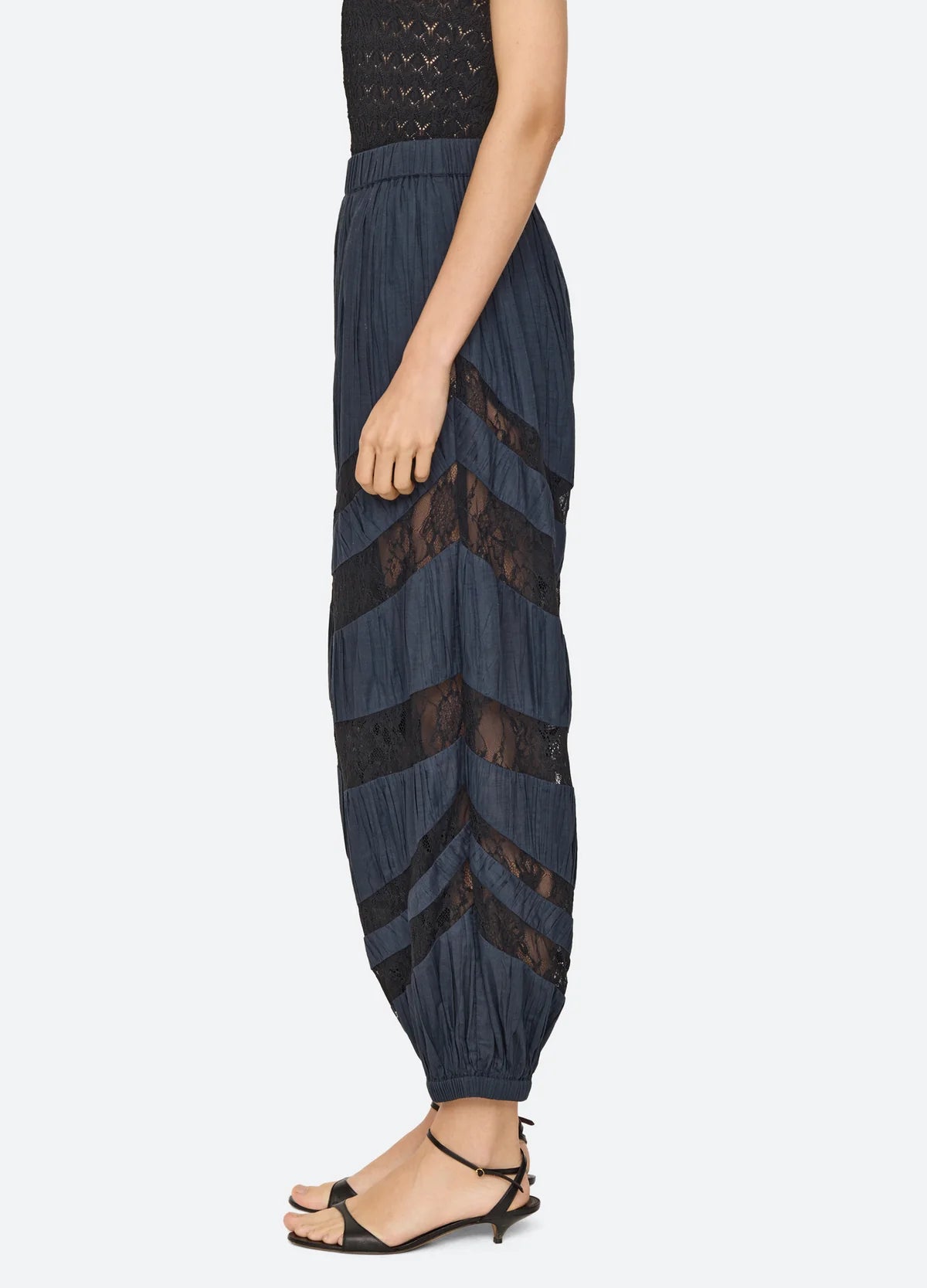 A side view of a model wearing high-waisted navy blue pleated harem-style pants with sheer black chevron lace panels, paired with black strappy sandals against a light gray background.