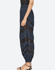 A side view of a model wearing high-waisted navy blue pleated harem-style pants with sheer black chevron lace panels, paired with black strappy sandals against a light gray background.