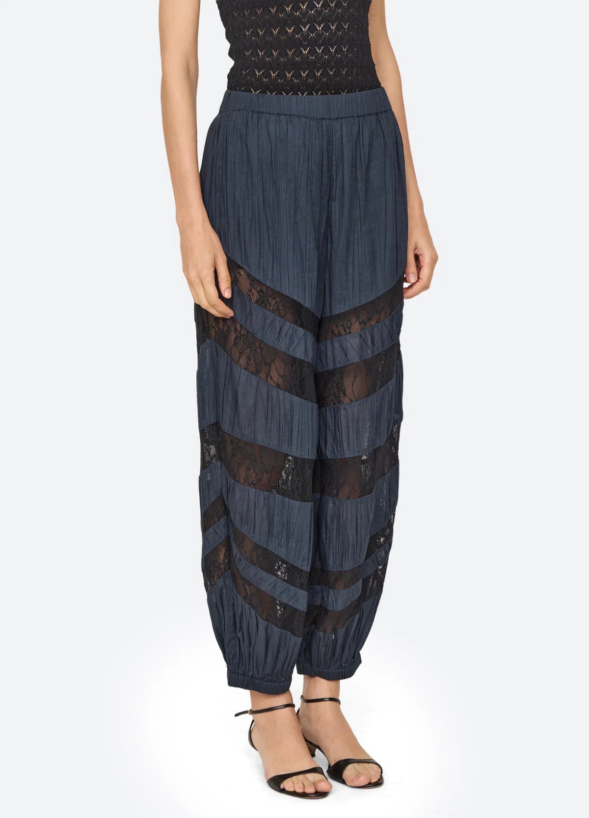 A model wearing high-waisted, voluminous navy blue pleated pants with sheer black lace horizontal bands, paired with a black top and black strappy sandals, against a light gray background.