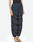 A model wearing high-waisted, voluminous navy blue pleated pants with sheer black lace horizontal bands, paired with a black top and black strappy sandals, against a light gray background.