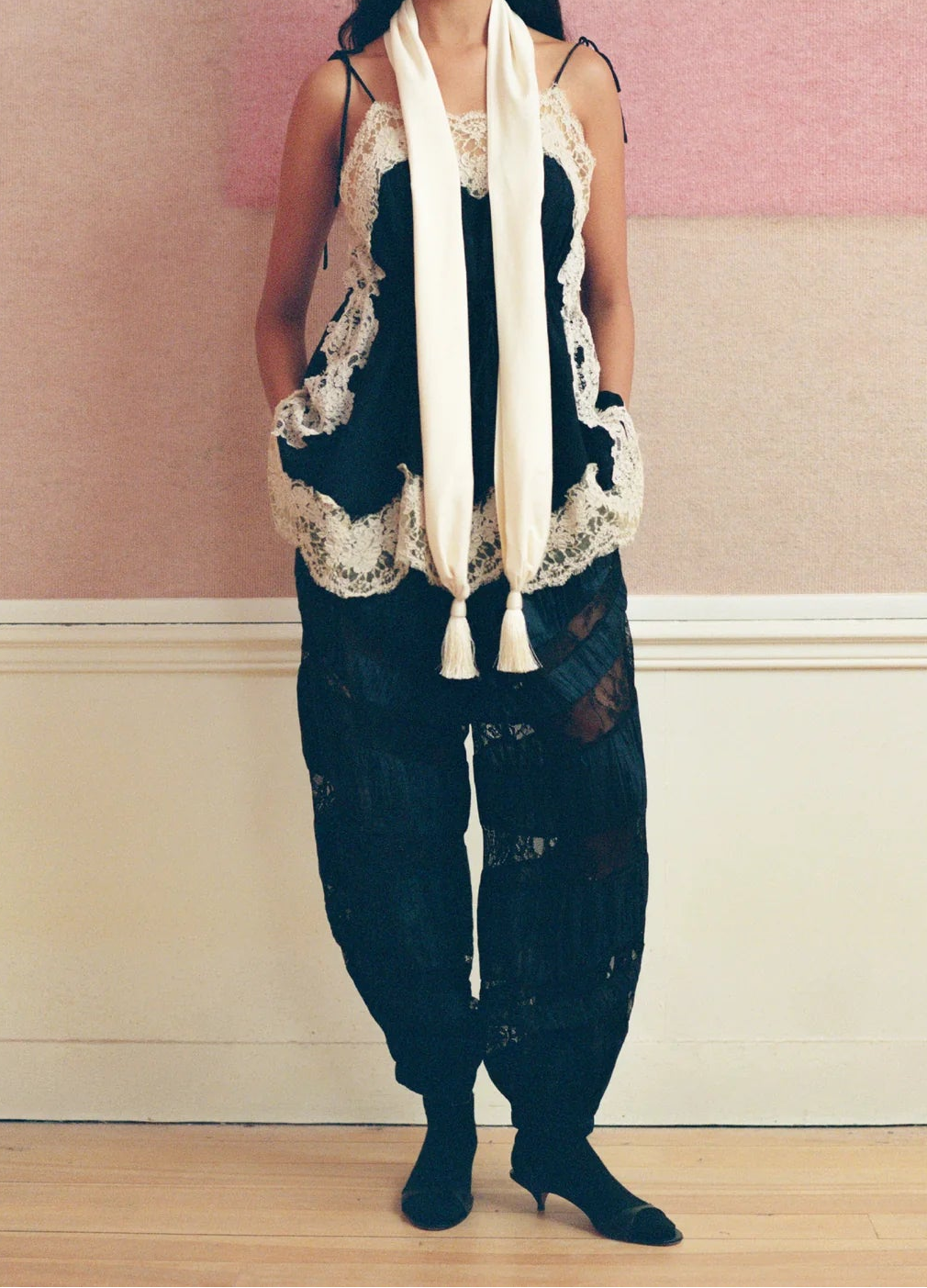 A model stands against a pink and cream wall wearing a black lace-trimmed camisole vest, navy and black lace pants, and a long cream scarf.