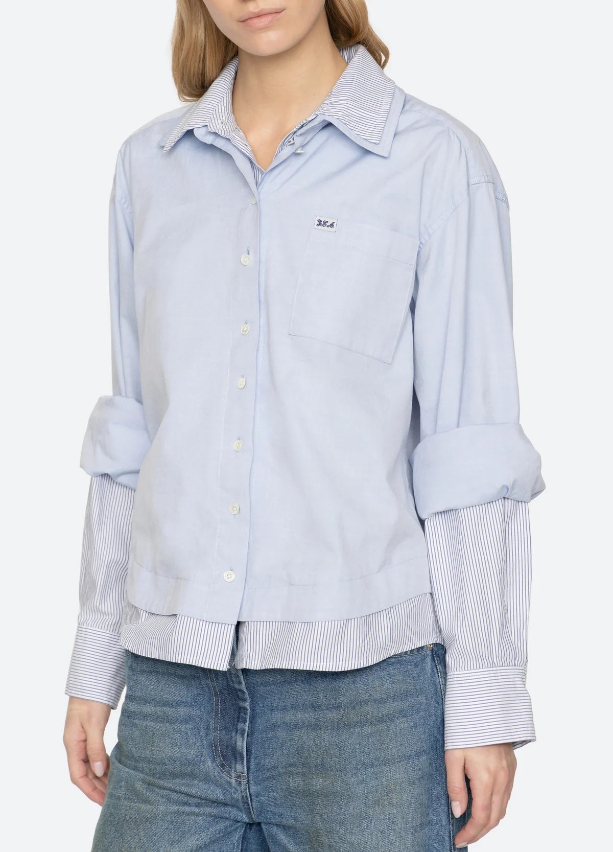 A woman wearing a light blue button-up shirt with a subtle textured weave, featuring a chest pocket, rolled-up sleeves, and a striped hem, paired with blue jeans against a plain white background.
