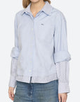 A woman wearing a light blue button-up shirt with a subtle textured weave, featuring a chest pocket, rolled-up sleeves, and a striped hem, paired with blue jeans against a plain white background.