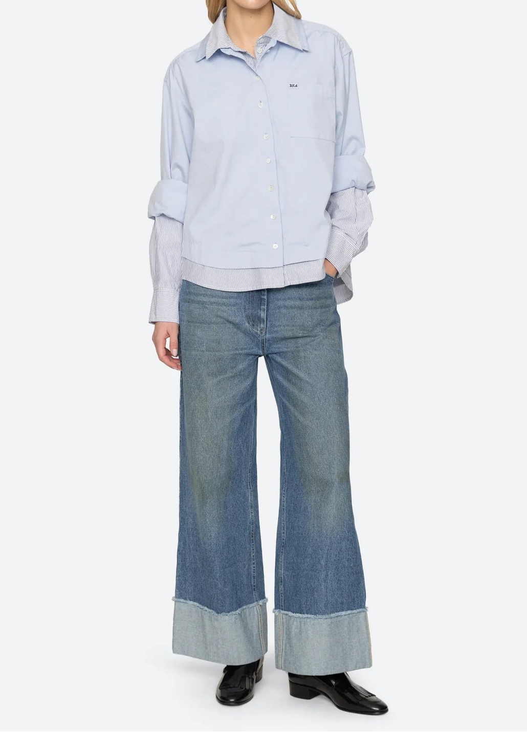 A model stands against a light gray studio background wearing a light blue button-up shirt layered over a white collared shirt, paired with wide-leg blue jeans featuring cuffed hems and black shoes.