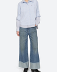 A model stands against a light gray studio background wearing a light blue button-up shirt layered over a white collared shirt, paired with wide-leg blue jeans featuring cuffed hems and black shoes.