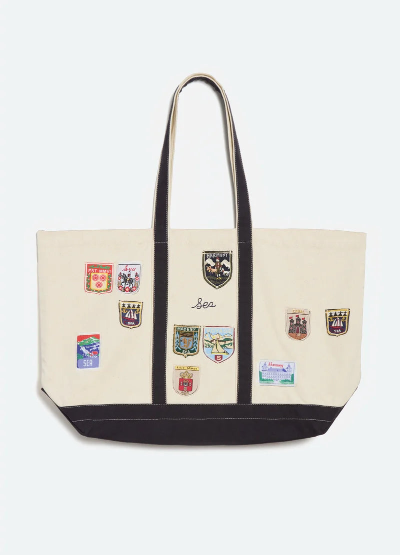 A cream canvas tote bag with black trim, dual shoulder straps, and multiple colorful embroidered school crest patches on the front, set against a plain light gray background.