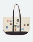 A cream canvas tote bag with black trim, dual shoulder straps, and multiple colorful embroidered school crest patches on the front, set against a plain light gray background.