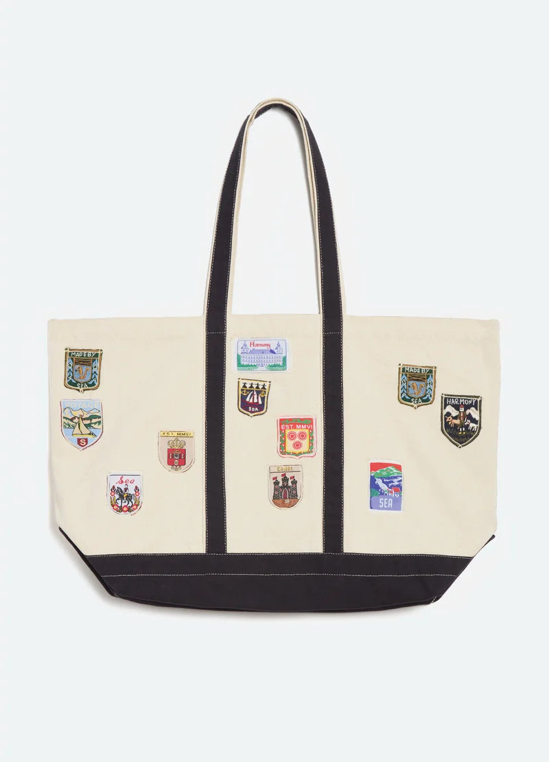 An off-white canvas tote bag with black trim, dual shoulder straps, and multiple colorful embroidered sports crests on the front, shown against a light gray background.
