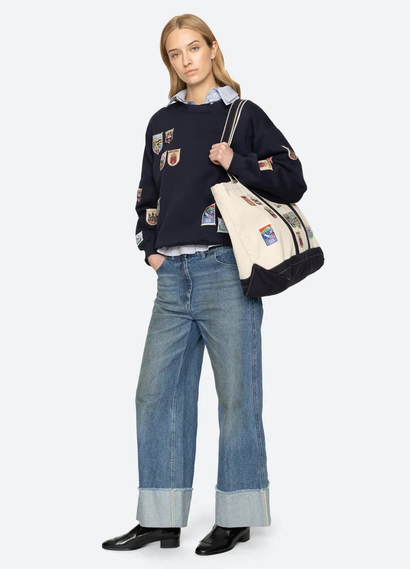 Person wearing a navy sweater with patches and blue jeans, holding a beige tote bag on a white background