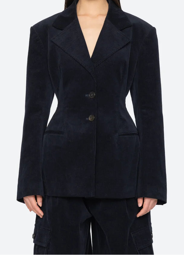 A model wearing a dark navy blue corduroy blazer with structured shoulders and a double-breasted button closure, paired with matching dark pants, against a light gray studio background.