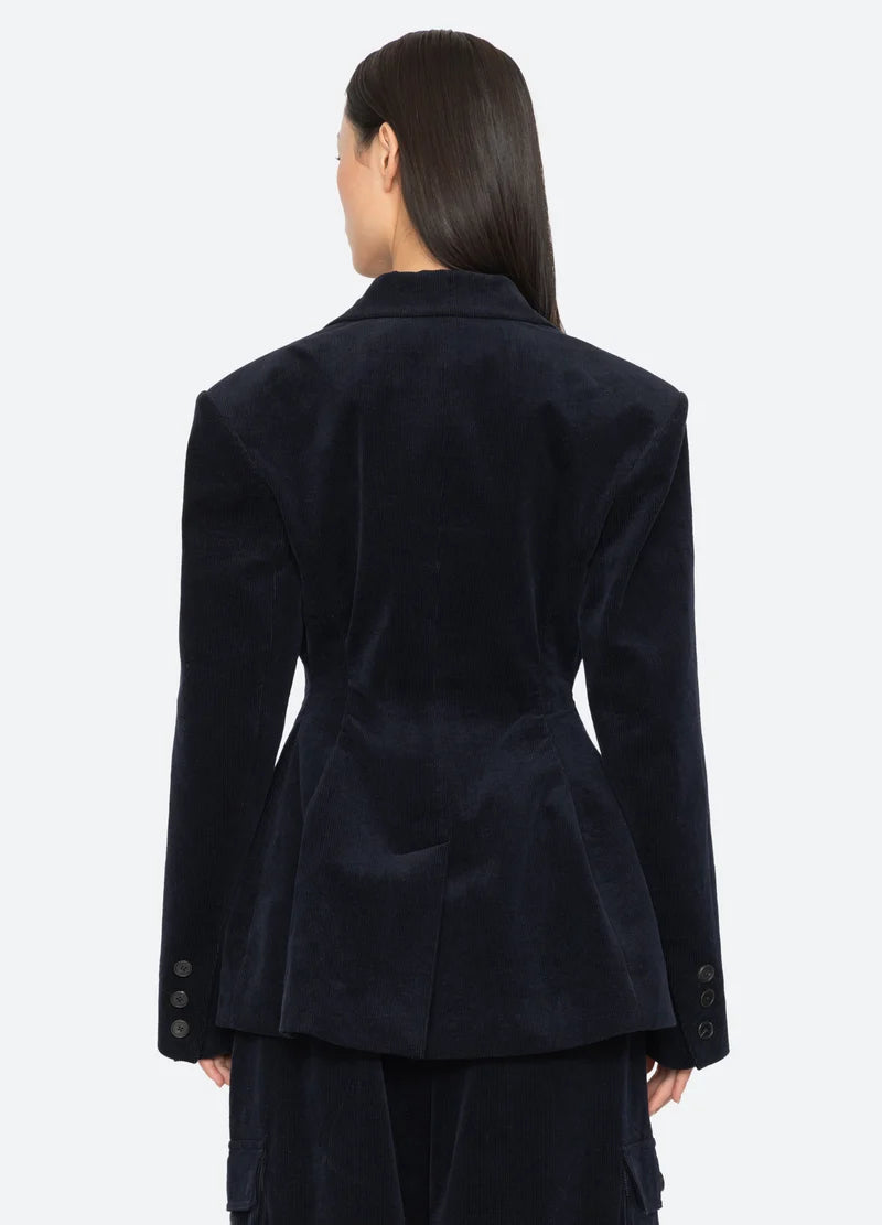 A model wearing a dark navy blue corduroy blazer paired with matching dark pants, against a light gray studio background, back view