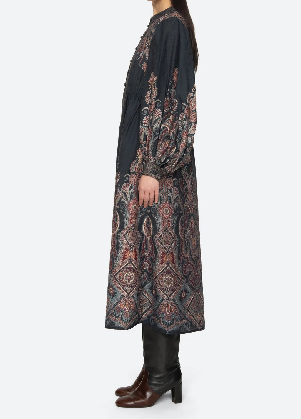 A model is shown from the side wearing a long-sleeved, dark charcoal dress with an intricate multicolor paisley and geometric pattern on the sleeves and skirt, paired with black leather boots against a light gray background.