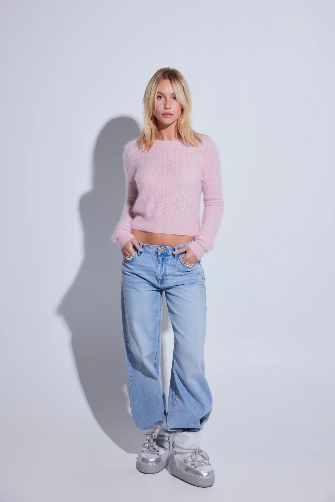 A woman stands against a light gray studio background wearing a fuzzy light pink cropped long-sleeve sweater, light blue wide-leg jeans, and metallic silver sneakers.