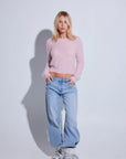 A woman stands against a light gray studio background wearing a fuzzy light pink cropped long-sleeve sweater, light blue wide-leg jeans, and metallic silver sneakers.