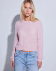 A blonde woman wearing a fuzzy light pink long-sleeve crop sweater and light blue jeans, standing against a plain light gray studio background.