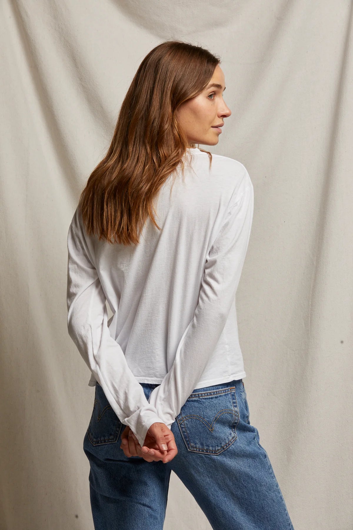 Woman wearing a white long-sleeve shirt and blue jeans against a plain background, back view, size S