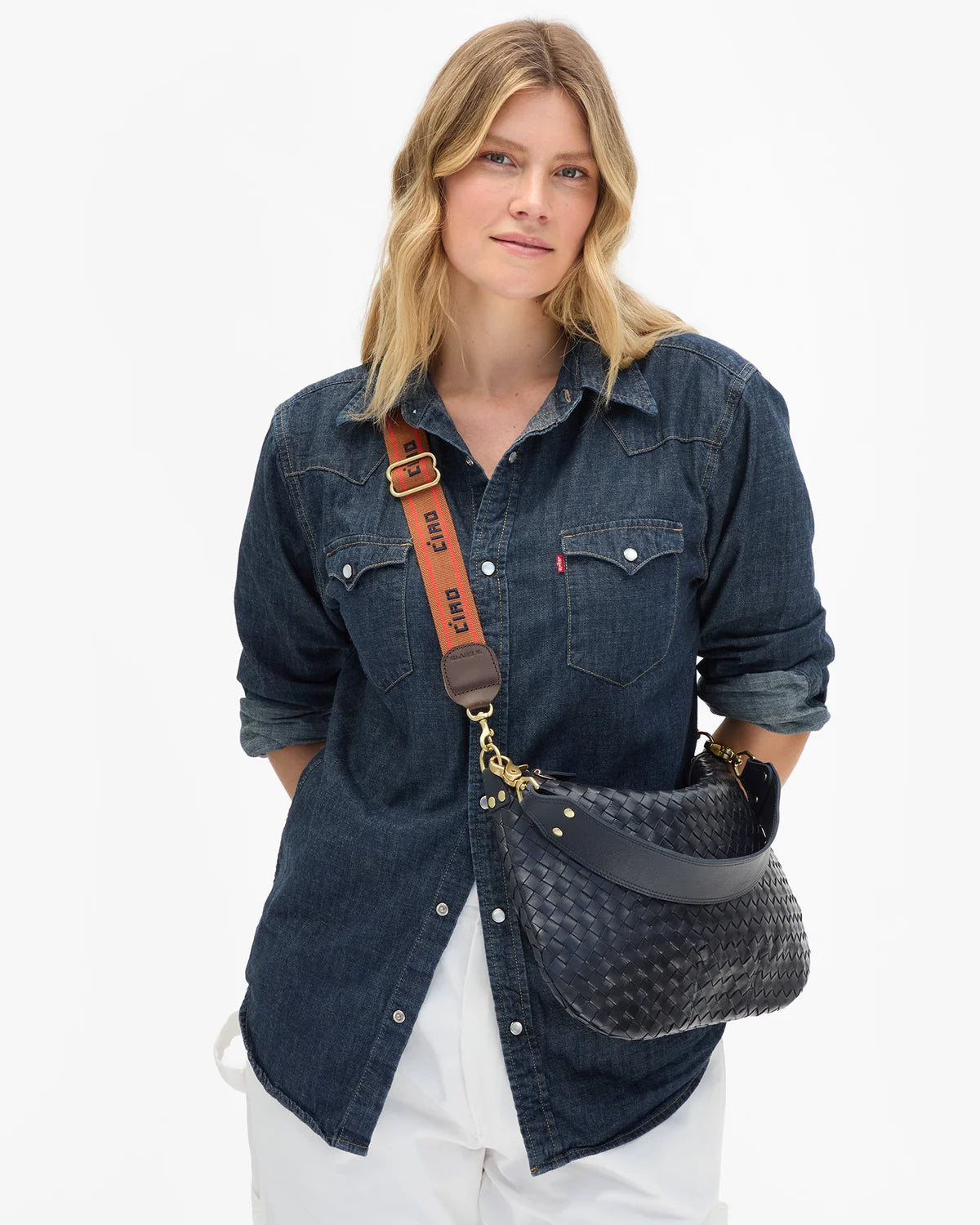 A woman wearing a dark blue denim shirt and white pants carries a dark woven shoulder bag with a brown strap featuring the brand name 'cvc'.