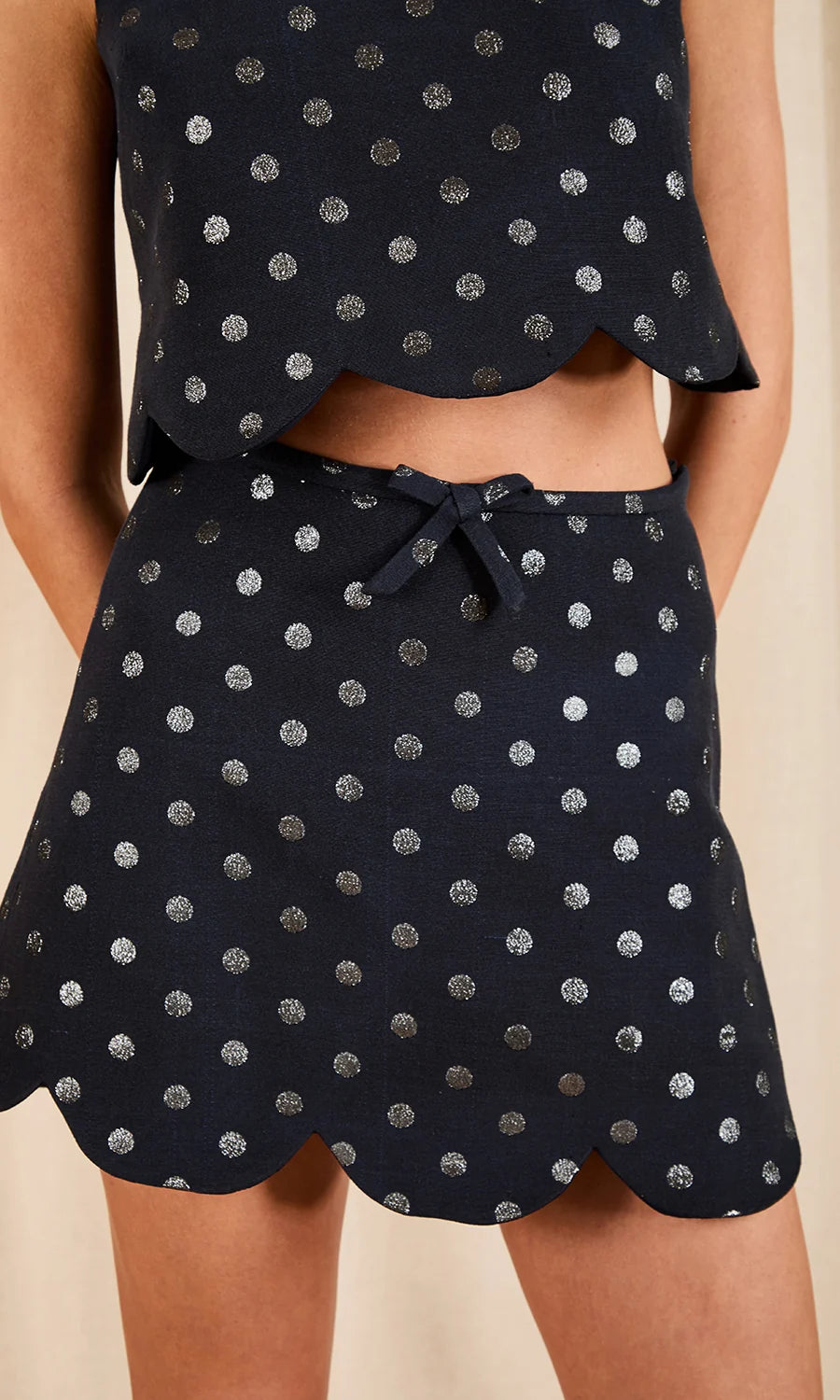 close up of a model wearing a dark colored top and skirt set with metallic polka dots on a cream background