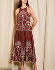 A woman modeling a sleeveless rust-brown maxi dress with white embroidered patterns, standing against a beige curtain backdrop.