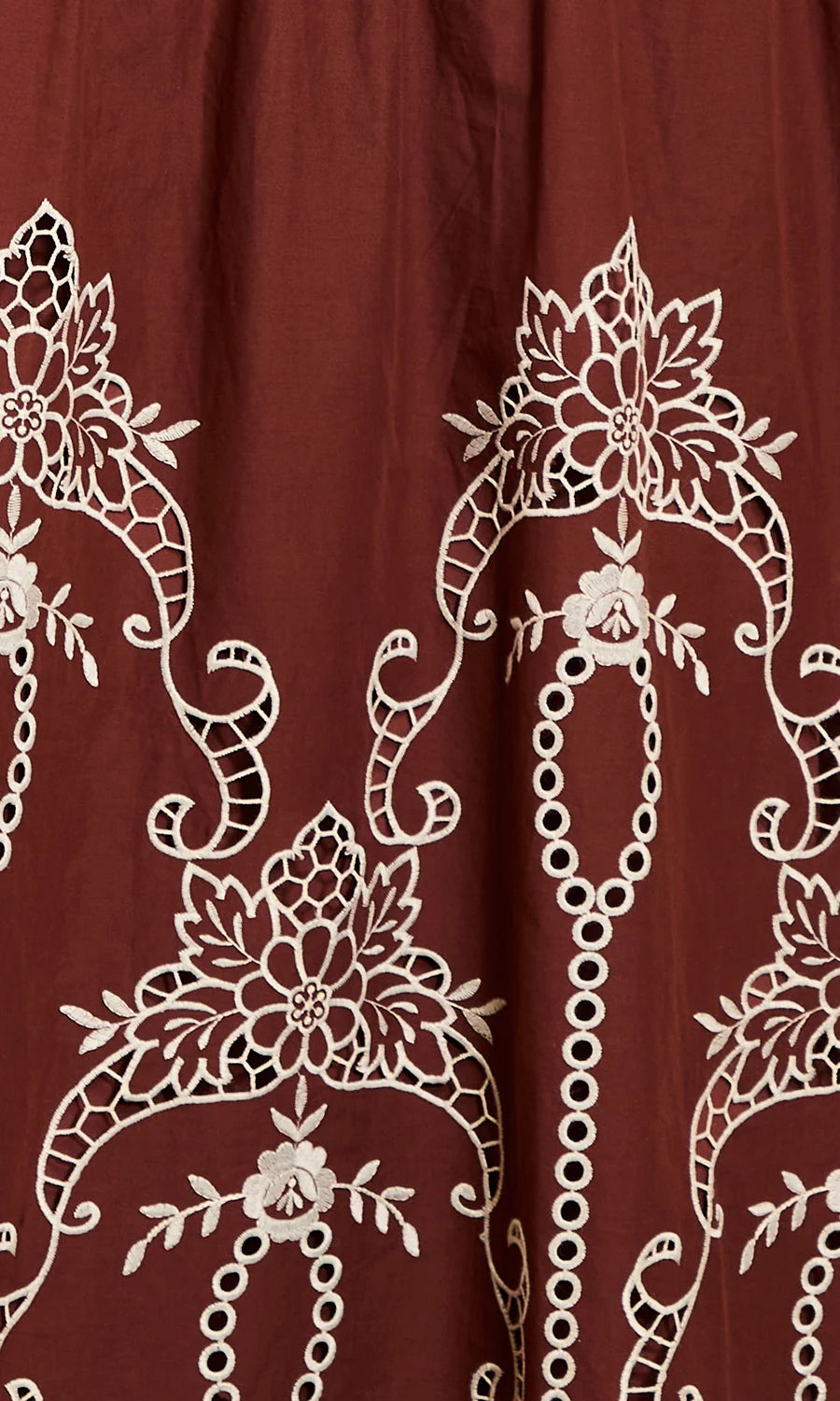 A close-up of a rust red or burgundy fabric with intricate white floral and scroll embroidery.