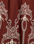 A close-up of a rust red or burgundy fabric with intricate white floral and scroll embroidery.