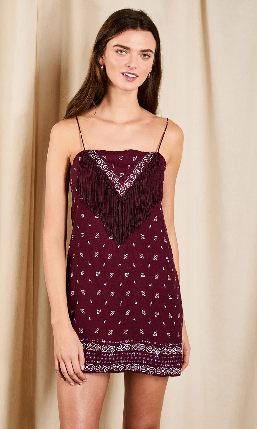 A woman modeling a burgundy mini dress with white geometric patterns and fringe detailing on the bodice, set against a neutral beige curtain backdrop.