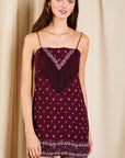 A woman modeling a burgundy mini dress with white geometric patterns and fringe detailing on the bodice, set against a neutral beige curtain backdrop.