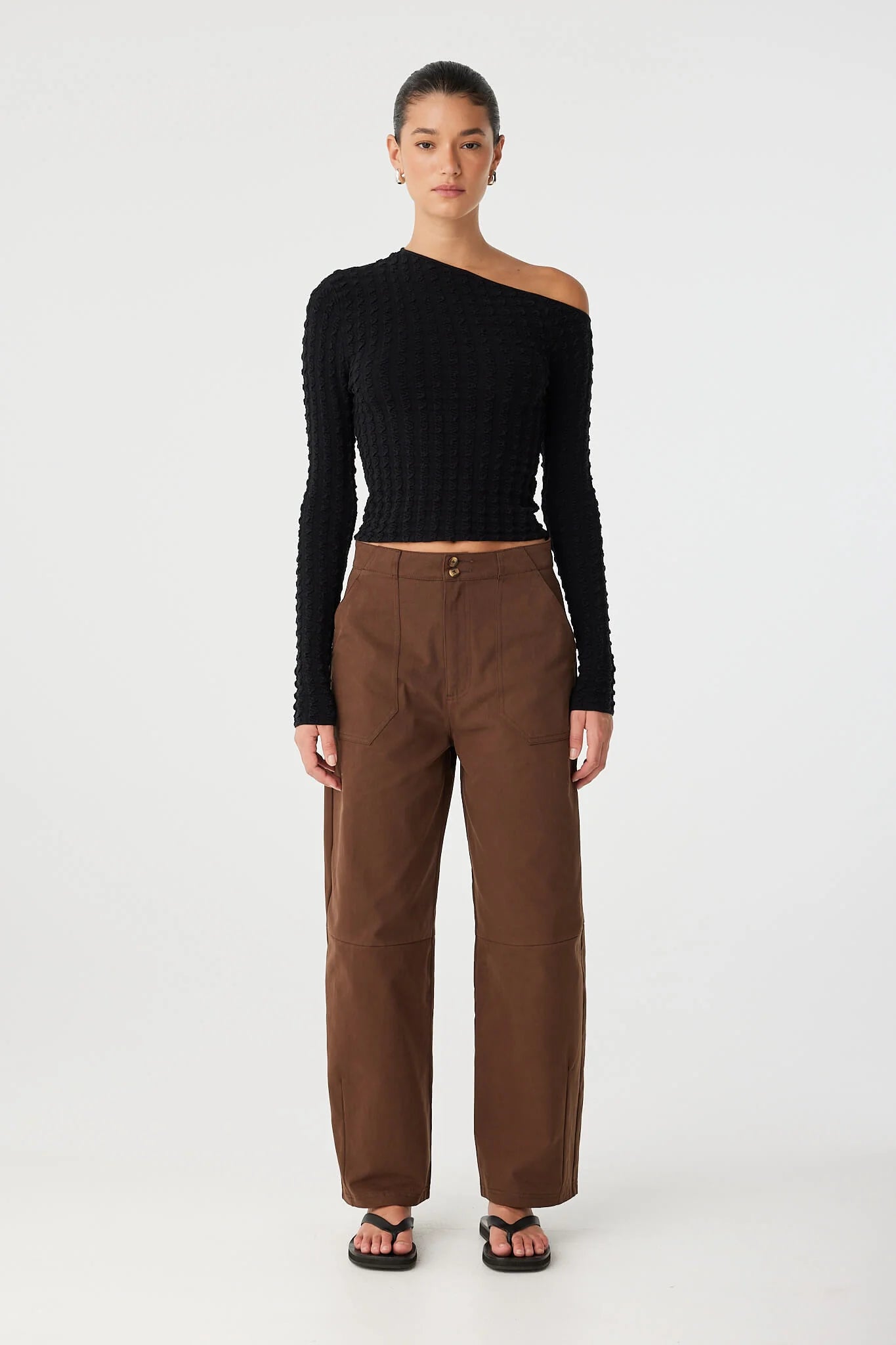 Woman wearing a black one-shoulder sweater and brown pants on a white background